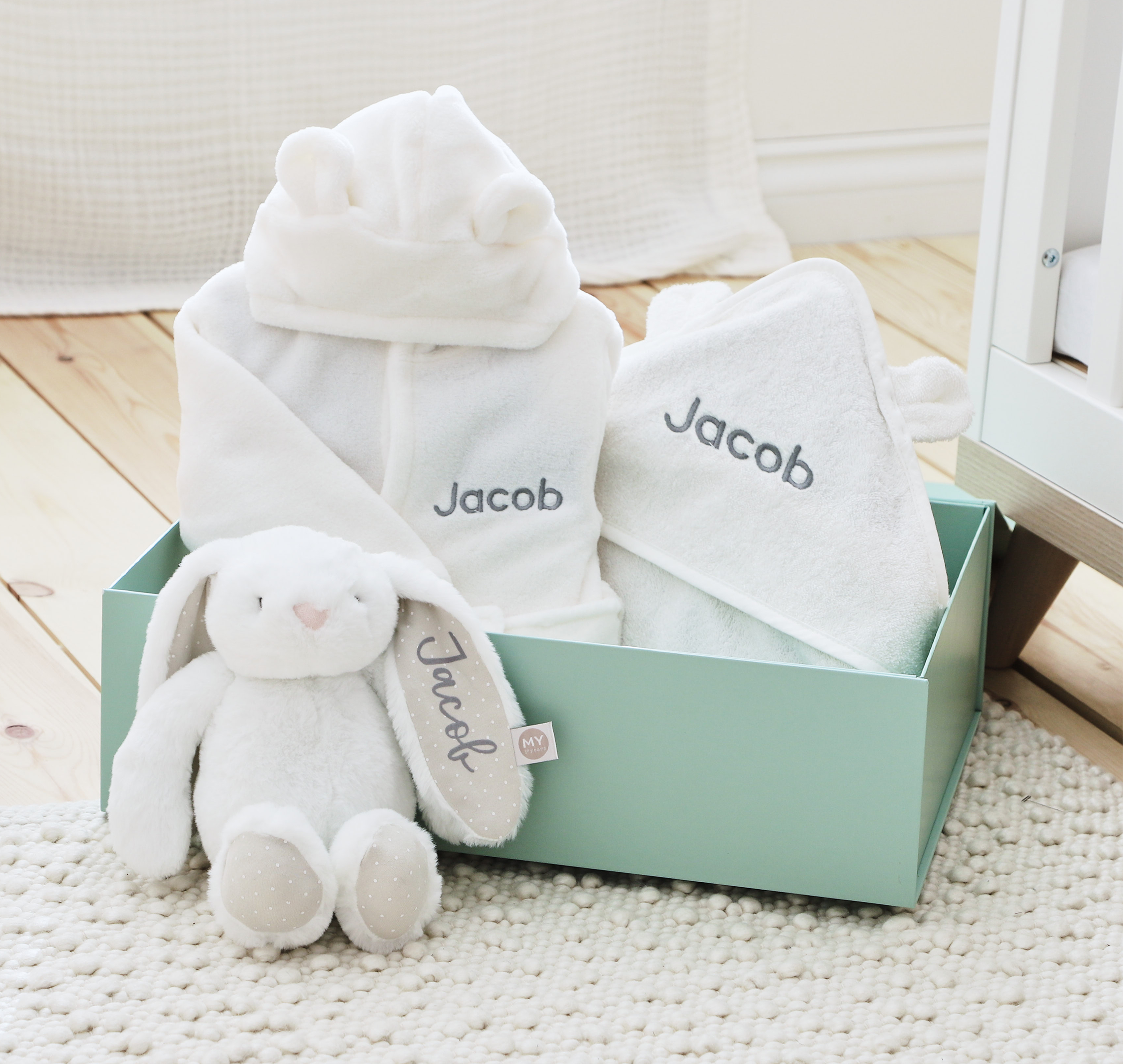 Soft personalised baby gift set with white hooded towels and cuddly bunny toy in a pastel green box on a nursery floor