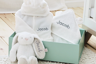 Soft personalised baby gift set with white hooded towels and cuddly bunny toy in a pastel green box on a nursery floor