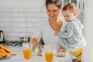 Eine Frau hält ein kleines Kind in einer Küche und bereitet eine Mahlzeit mit verschiedenen Lebensmitteln auf der Theke vor.