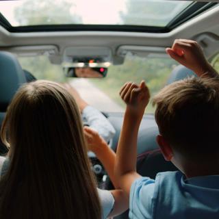 Zwei Personen, vermutlich ein Kind und ein Erwachsener, in einem Auto, wobei das Kind die Hand hebt und die Fahrt zu genießen scheint.