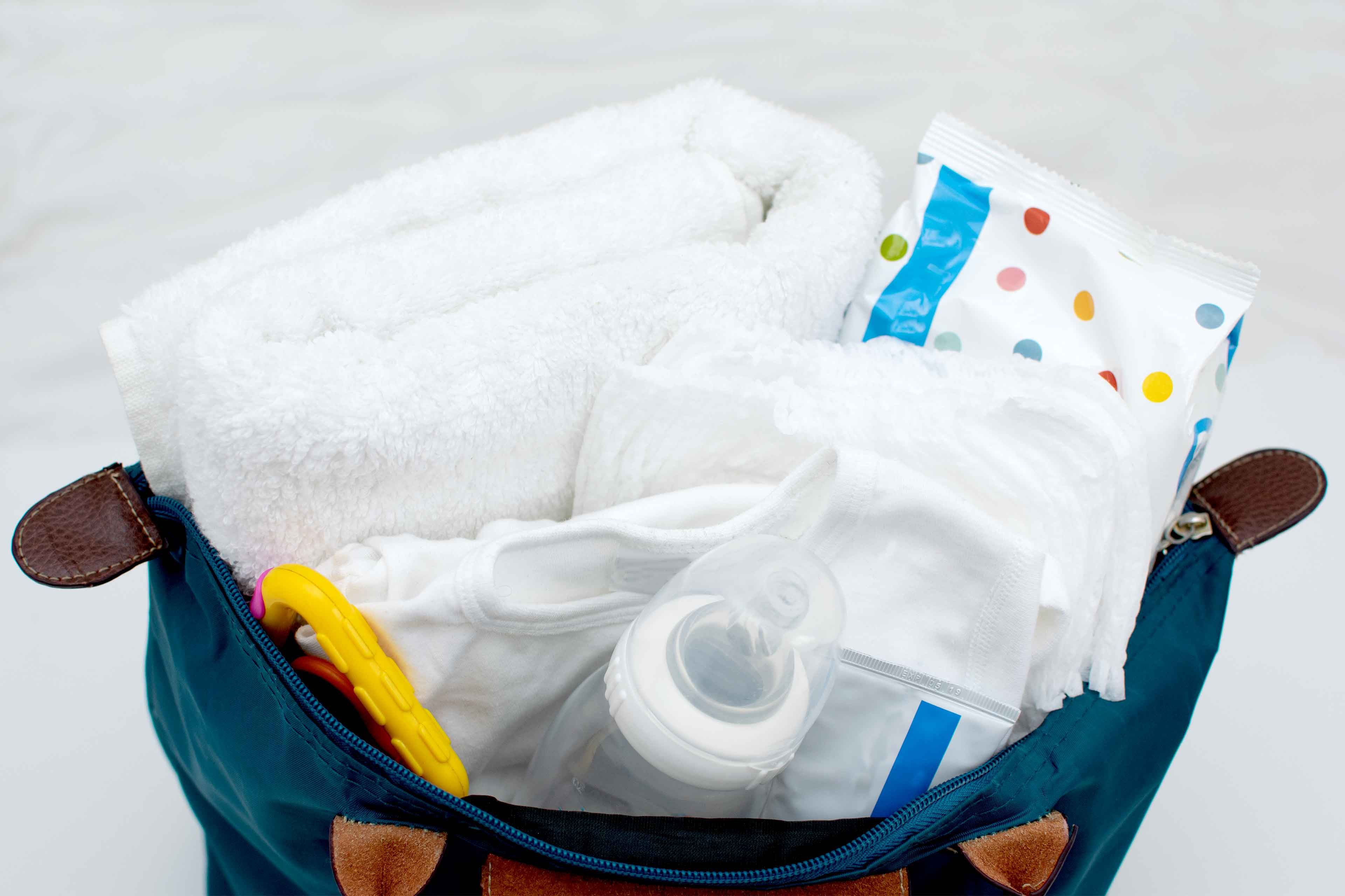 Eine blaue Tasche mit Babyutensilien wie einem weißen Handtuch, einer Babyflasche, einem Schnuller und einem bunten Beißring.