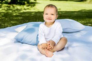 Ein fröhliches Baby, das auf einer weißen Decke im Freien sitzt, umgeben von Grünzeug und blauen Kissen.