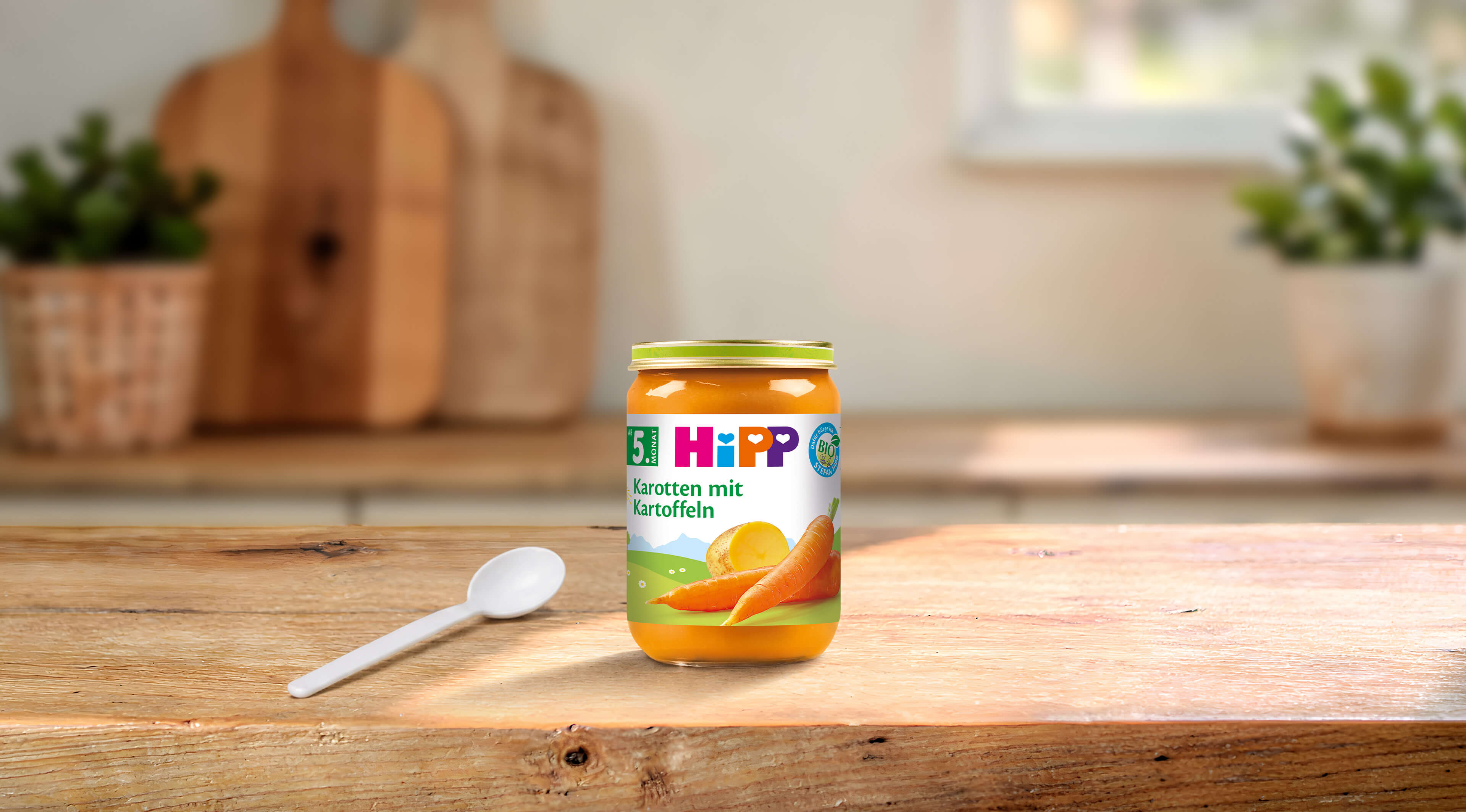 HiPP Bio Babygläschen Karotten mit Kartoffeln auf Holztisch mit Löffel in heller, gemütlicher Küche