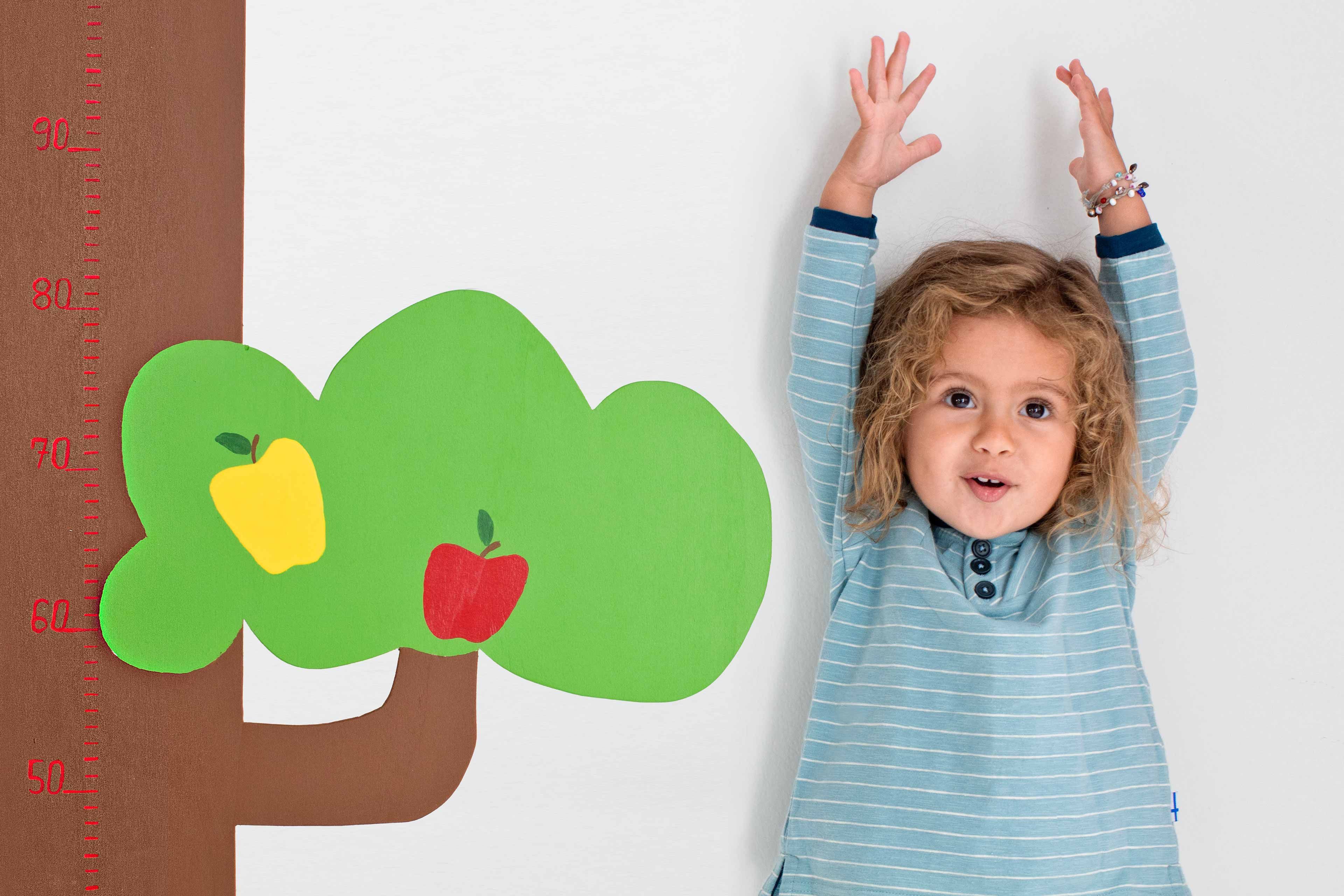 Ein kleines Kind mit lockigem Haar, das ein blau gestreiftes Hemd trägt und sich mit beiden Händen an einer Wand abstützt, neben einem grünen Papierbaum mit Äpfeln.