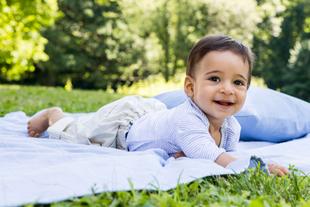 Ein fröhliches Baby, das auf einer blauen Decke auf einer Wiese liegt und ein blaues Kissen neben sich hat.