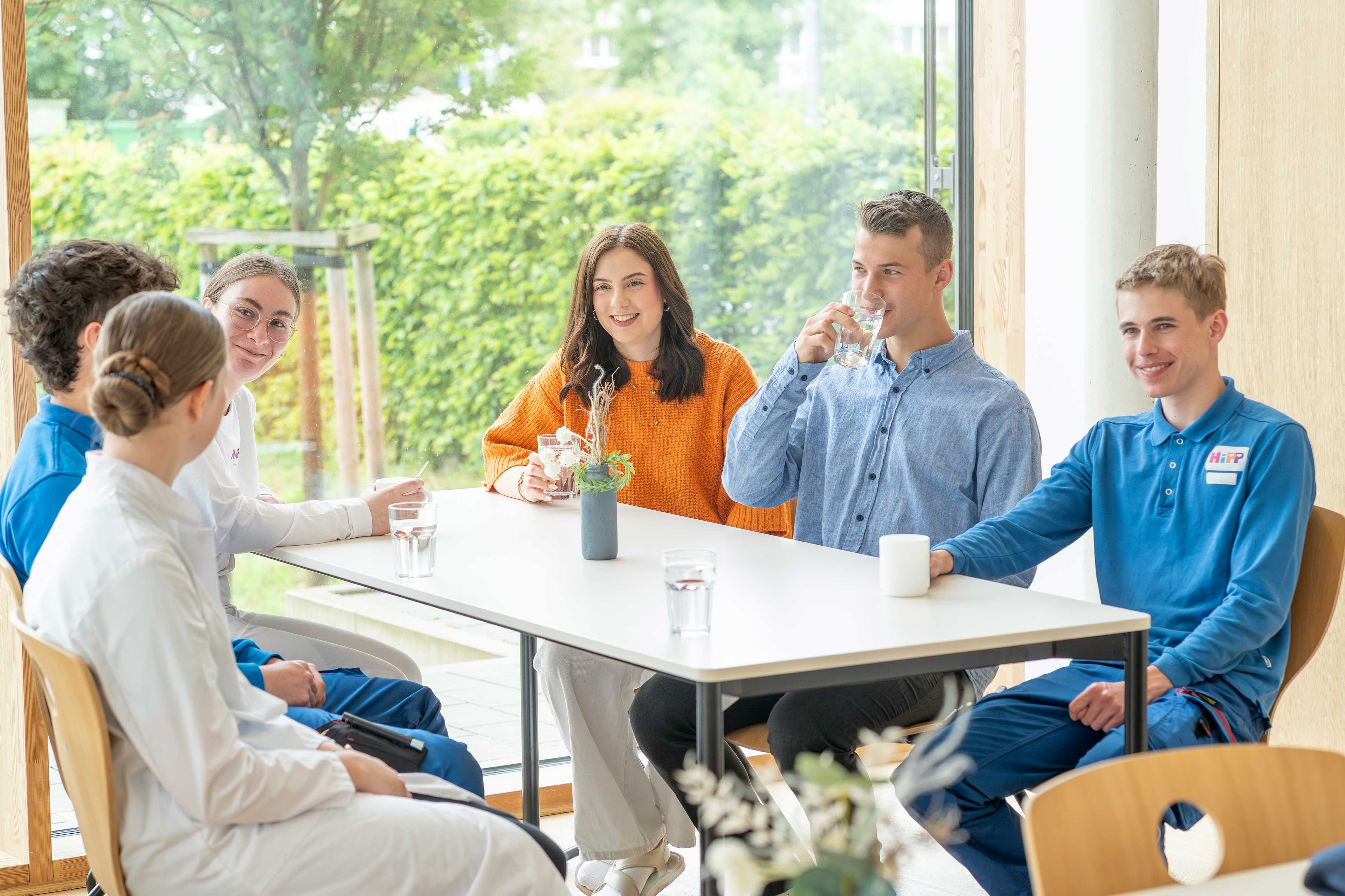 Junge HiPP Mitarbeitende und Auszubildende sitzen entspannt gemeinsam am Tisch und unterhalten sich in einer hellen Kantine.