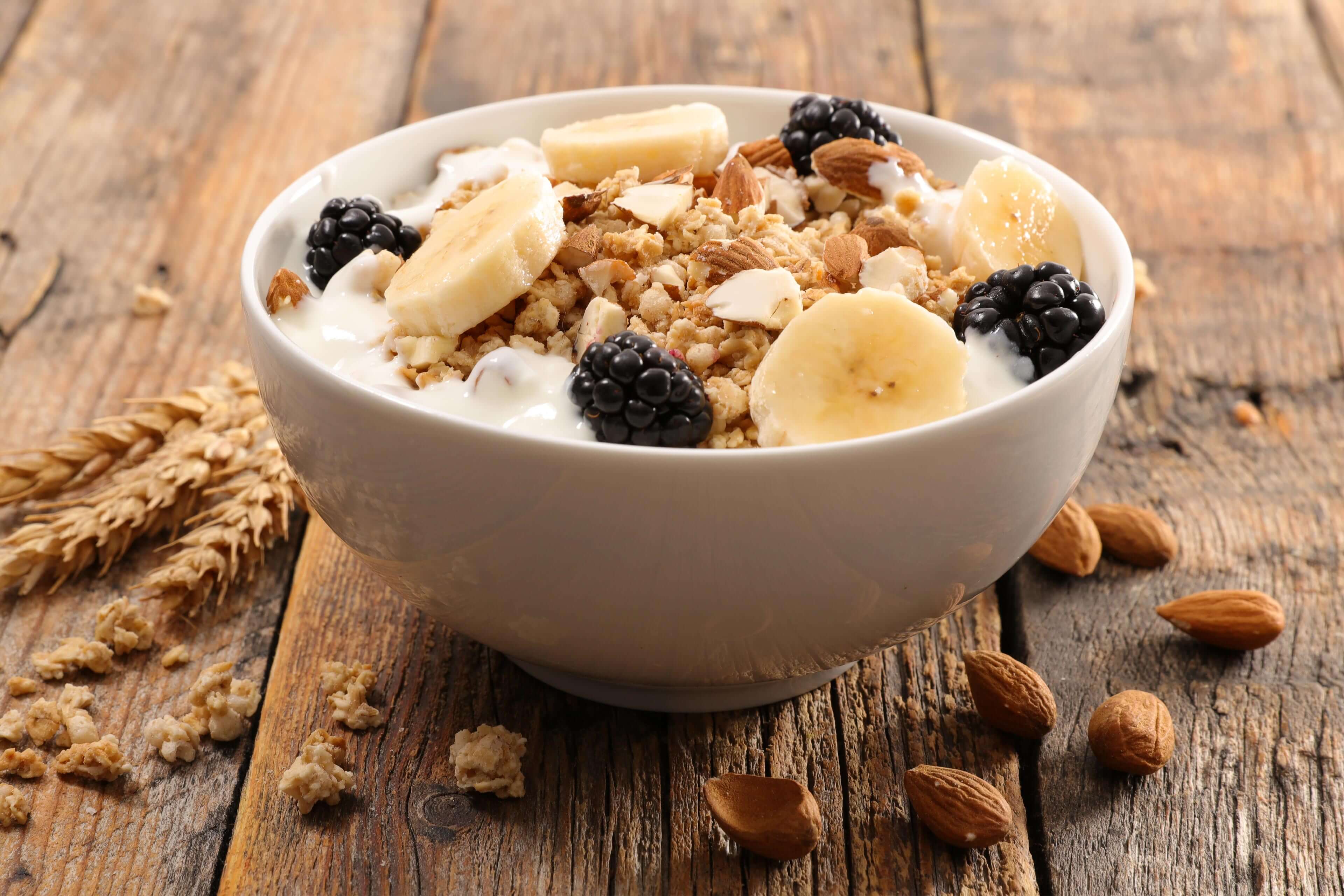Eine Schale Frühstücksflocken mit Bananenscheiben, Brombeeren und Mandeln, begleitet von einem Weizenstängel und Mandeln auf einer rustikalen Holzunterlage.