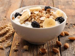 Eine Schale Frühstücksflocken mit Bananenscheiben, Brombeeren und Mandeln, begleitet von einem Weizenstängel und Mandeln auf einer rustikalen Holzunterlage.