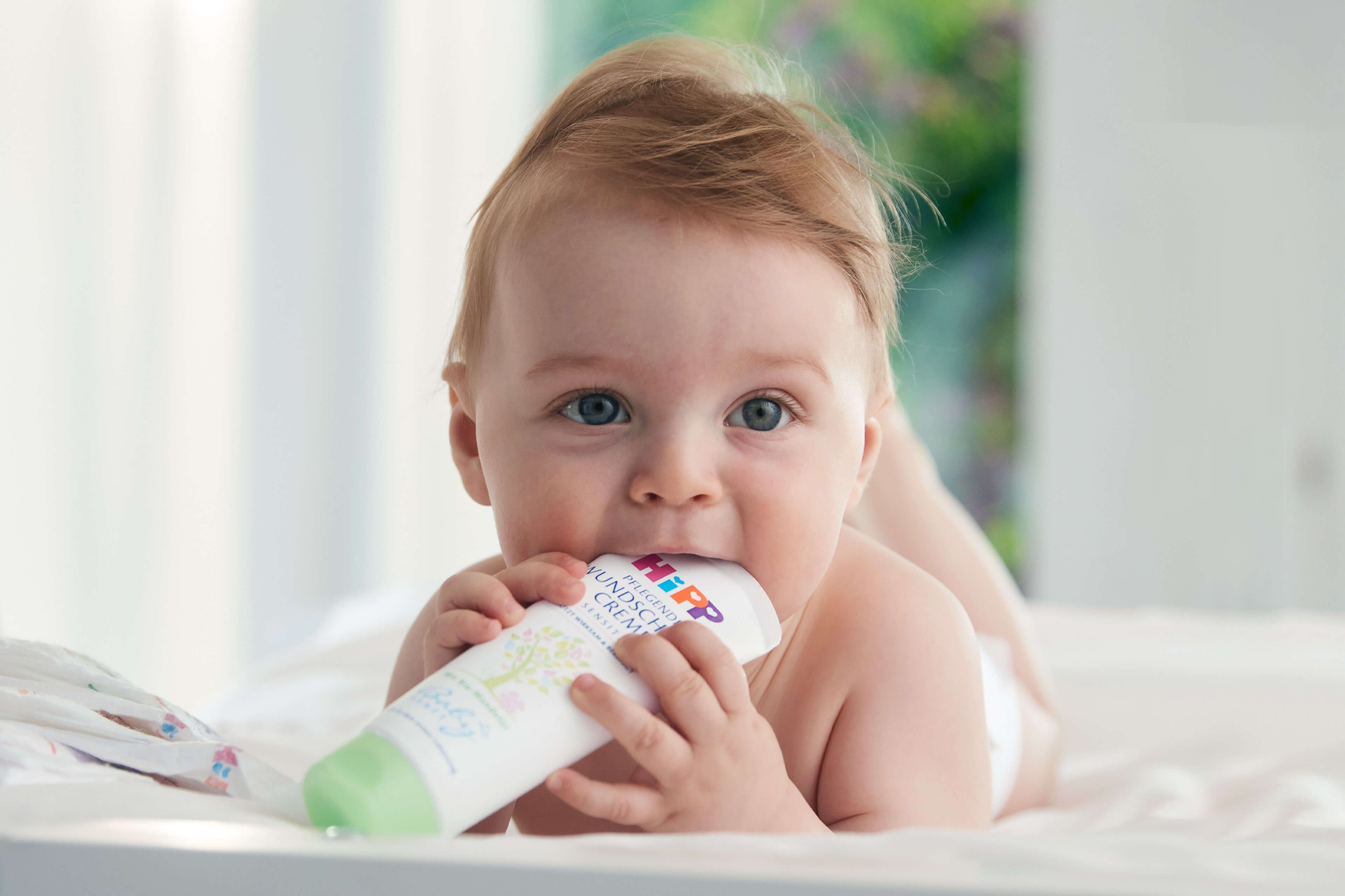 Ein Baby mit blauen Augen, das auf einem Bett liegt und eine Tube Babylotion mit der Aufschrift "Baby's Unscented" hält.