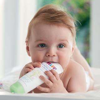 Ein Baby mit blauen Augen, das auf einem Bett liegt und eine Tube Babylotion mit der Aufschrift "Baby's Unscented" hält.