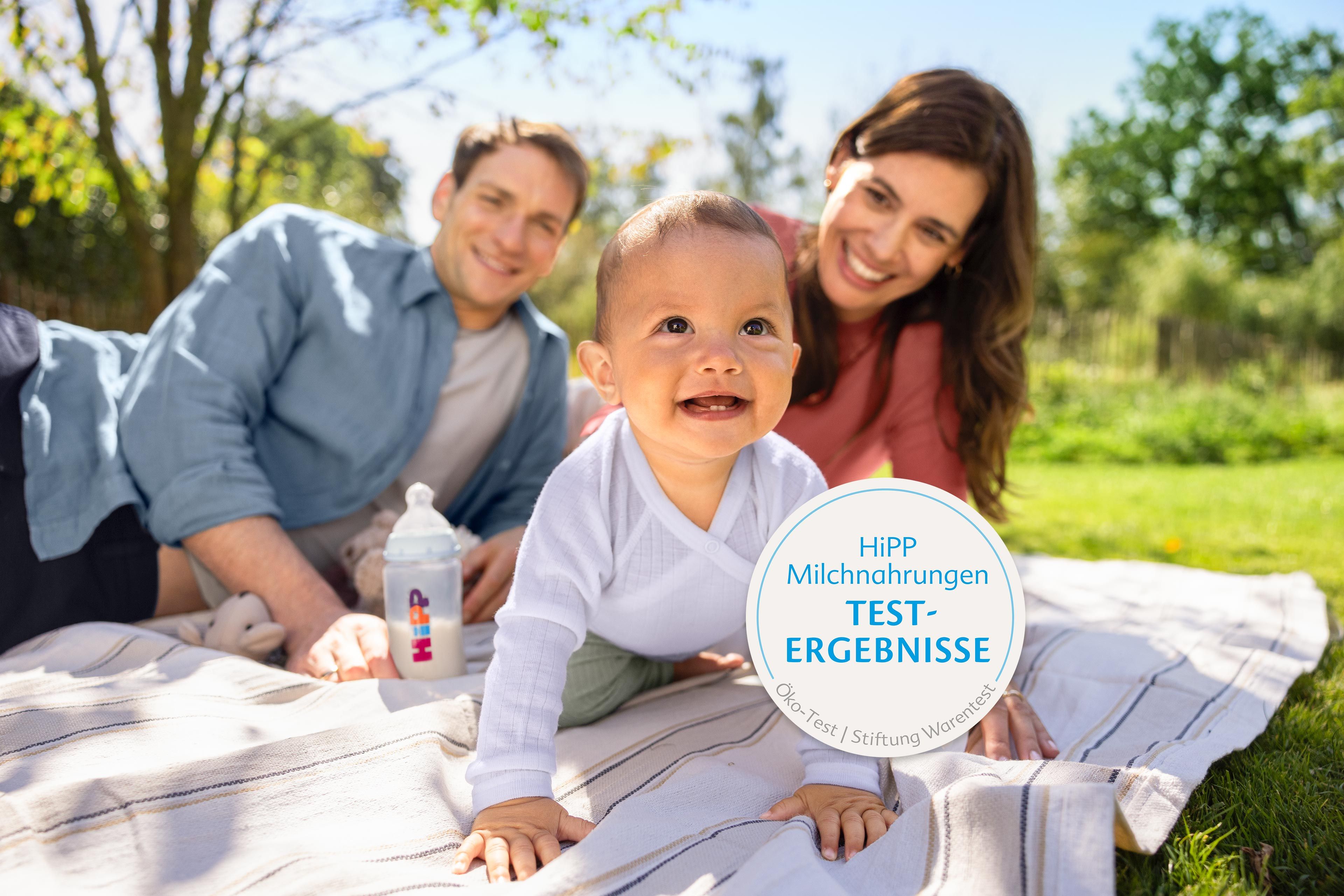 Lachendes Baby krabbelt auf einer Picknickdecke zwischen seinen Eltern, daneben eine HiPP-Flasche und ein Siegel zu Testergebnissen.