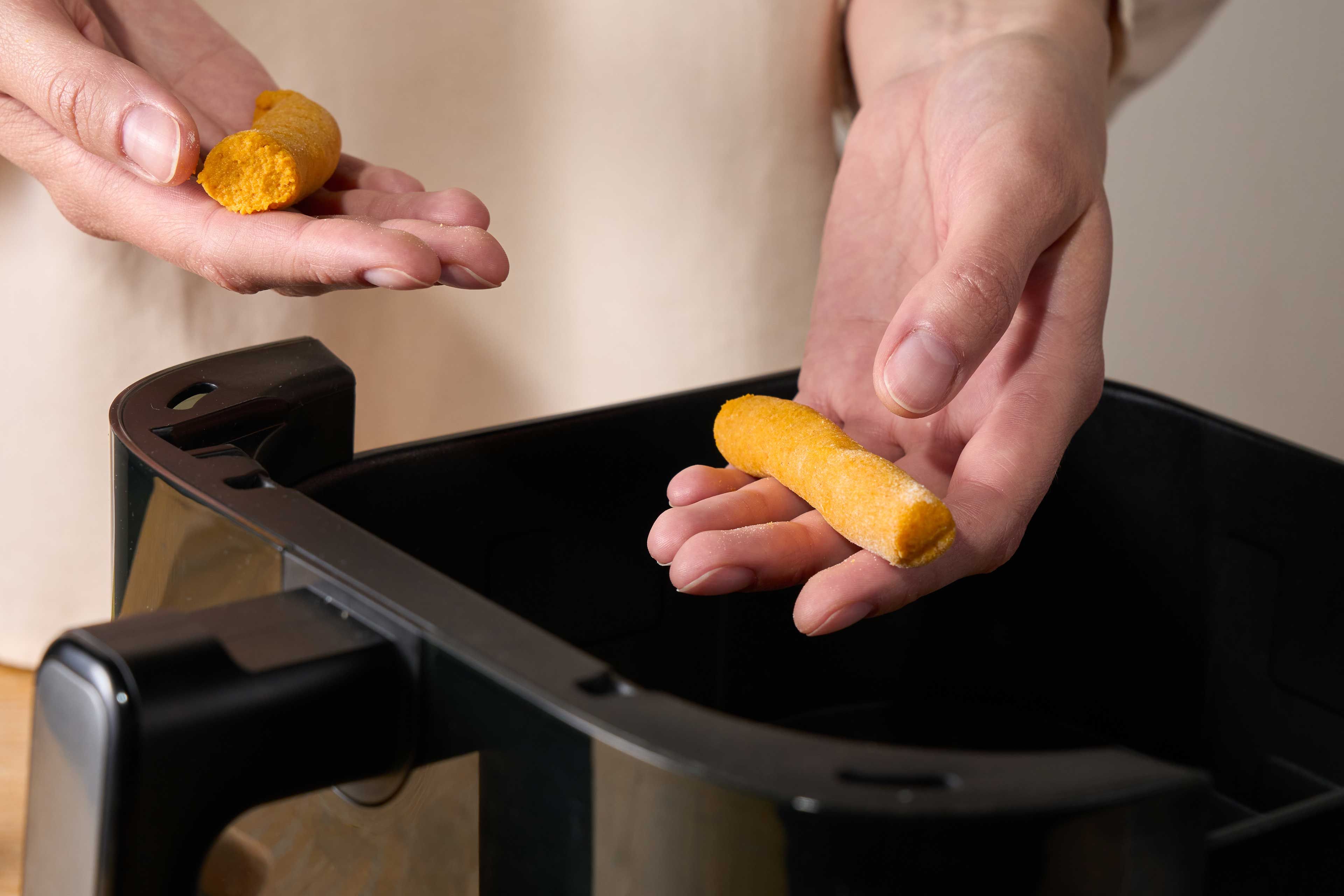 Hände halten zwei unterschiedlich geformte, gelbe Teigstücke über einem schwarzen Frittierkorb