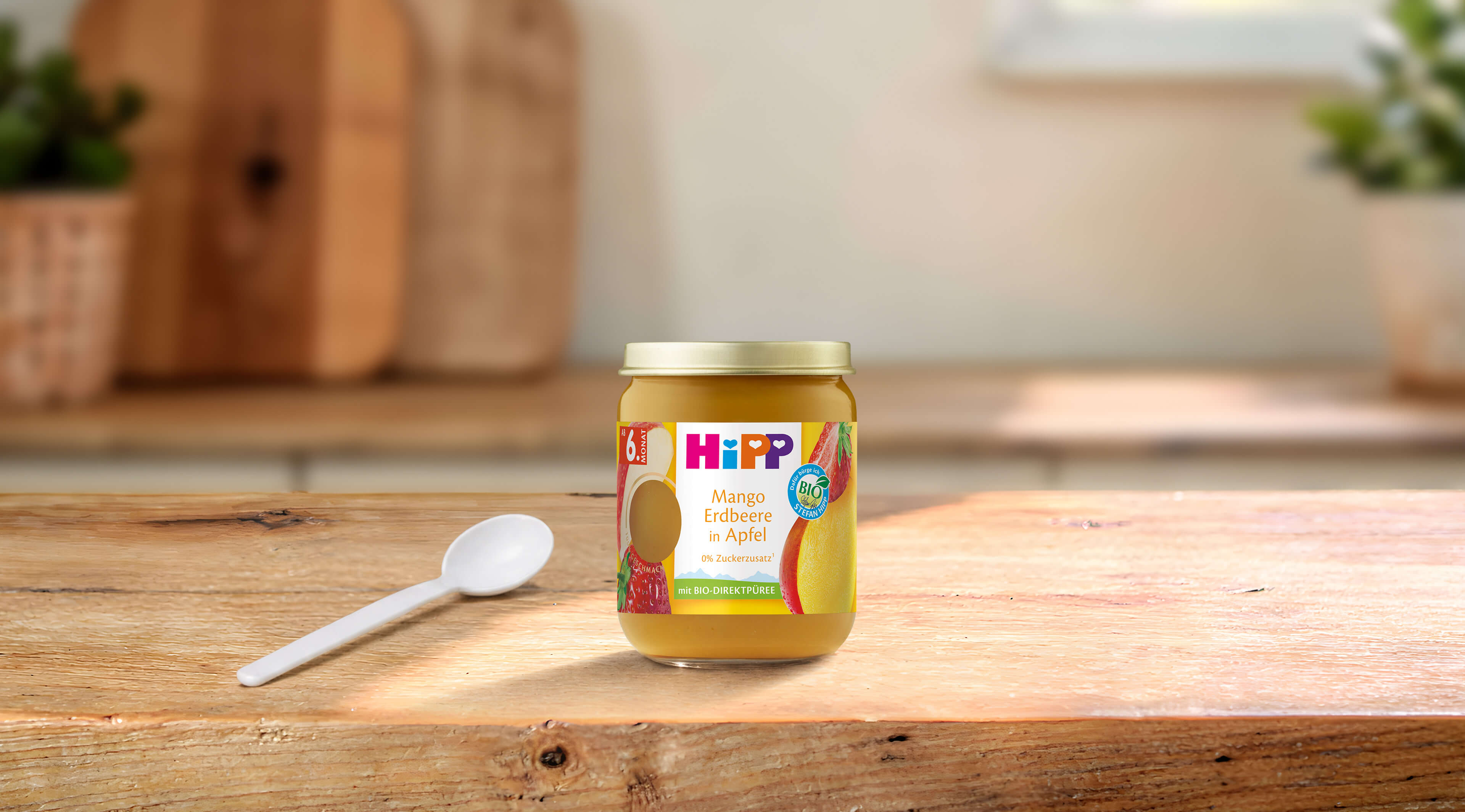 HiPP Bio-Früchte-Gläschen Mango Erdbeere in Apfel ab 6 Monaten auf Holztisch mit Löffel in heller Küche