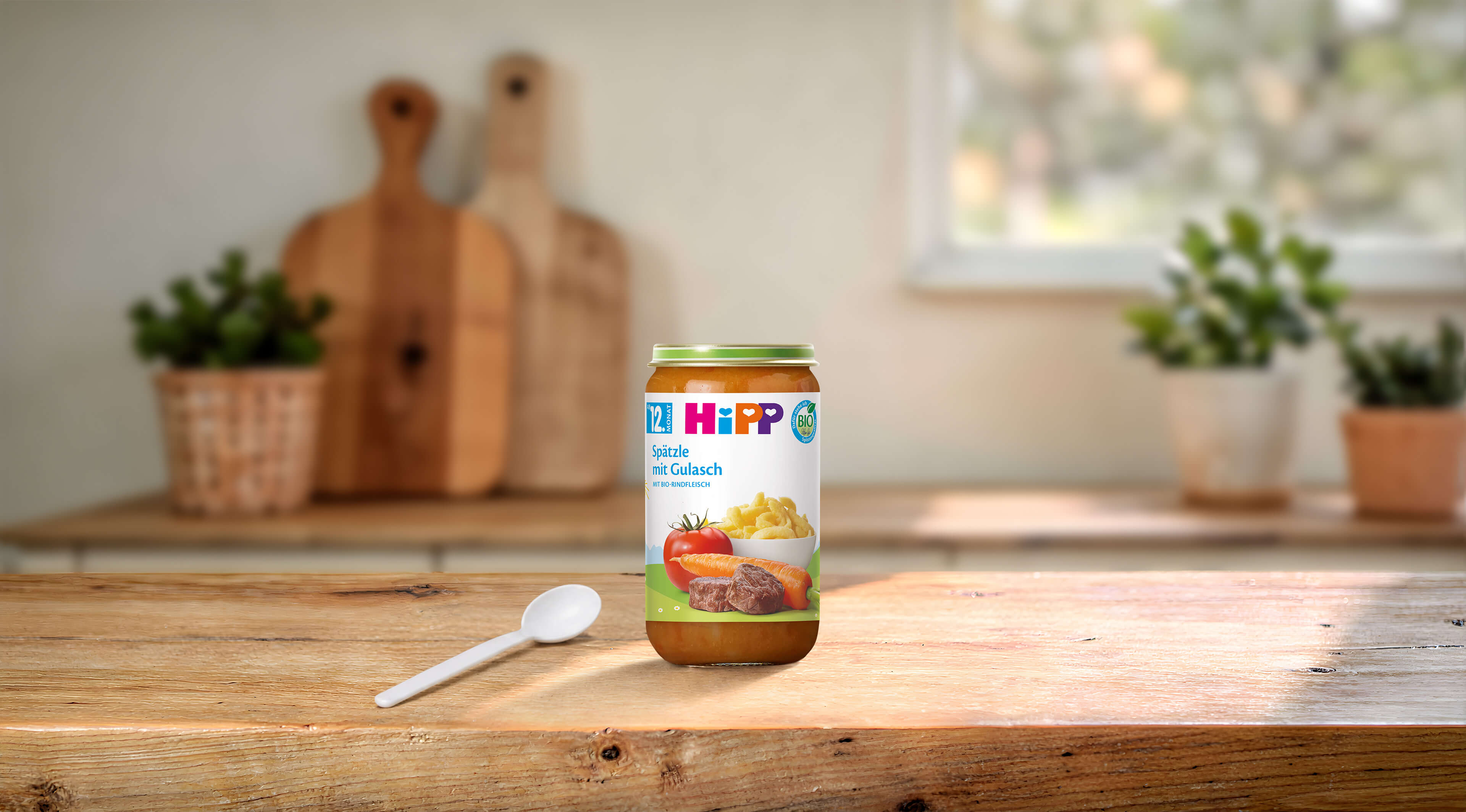 HiPP Bio Babygläschen Spätzle mit Gulasch 250g auf Holztisch, daneben ein Löffel in heller Küchenumgebung