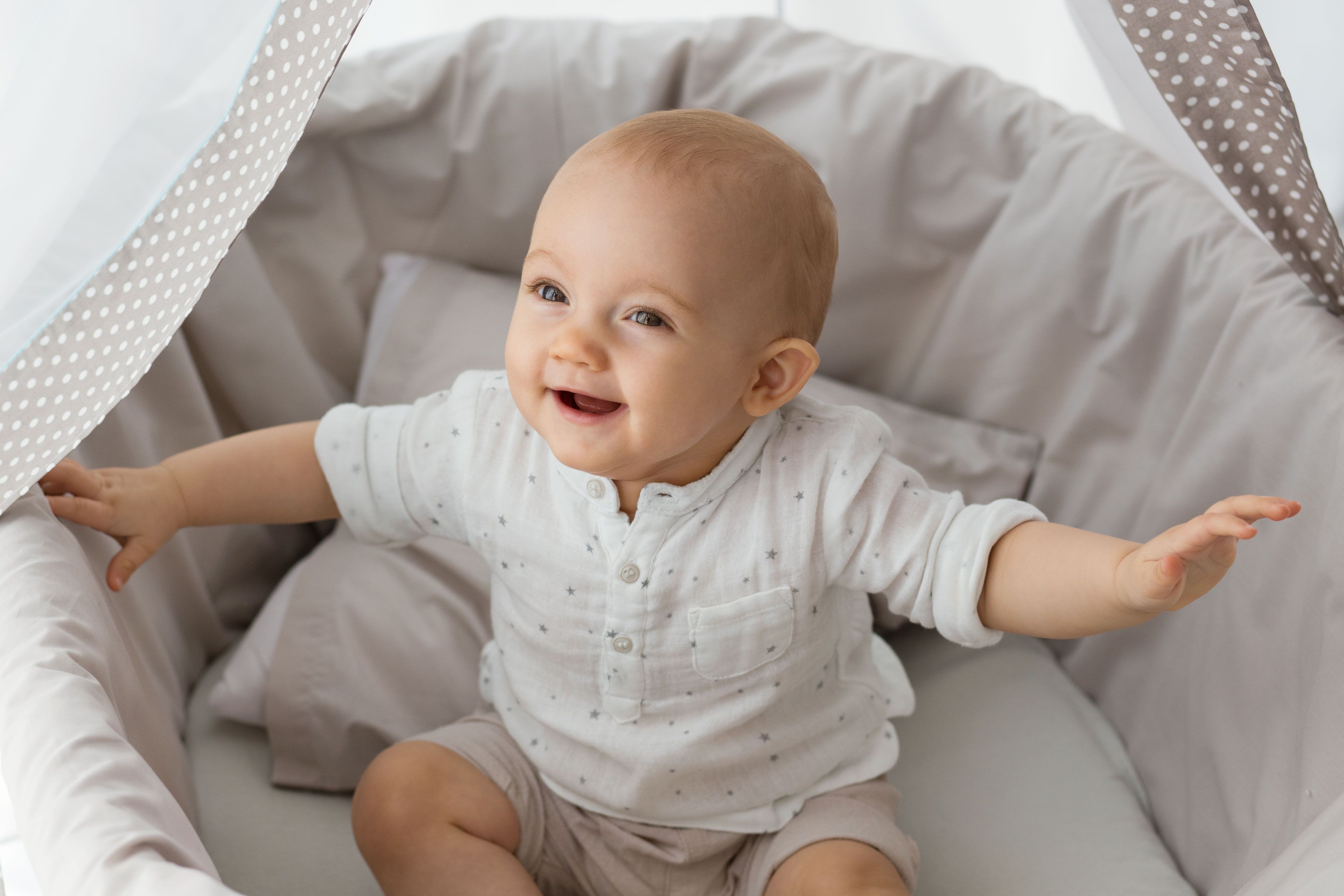 Ein Baby, bekleidet mit einem weißen Kleid mit kleinen Mustern, sitzt in einem Kinderbett mit gepunktetem Himmel.