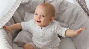 Ein Baby, bekleidet mit einem weißen Kleid mit kleinen Mustern, sitzt in einem Kinderbett mit gepunktetem Himmel.