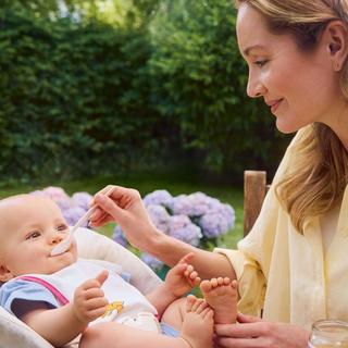 Frau in hellem Hemd füttert Baby im Hochstuhl im Garten mit Blumen im Hintergrund