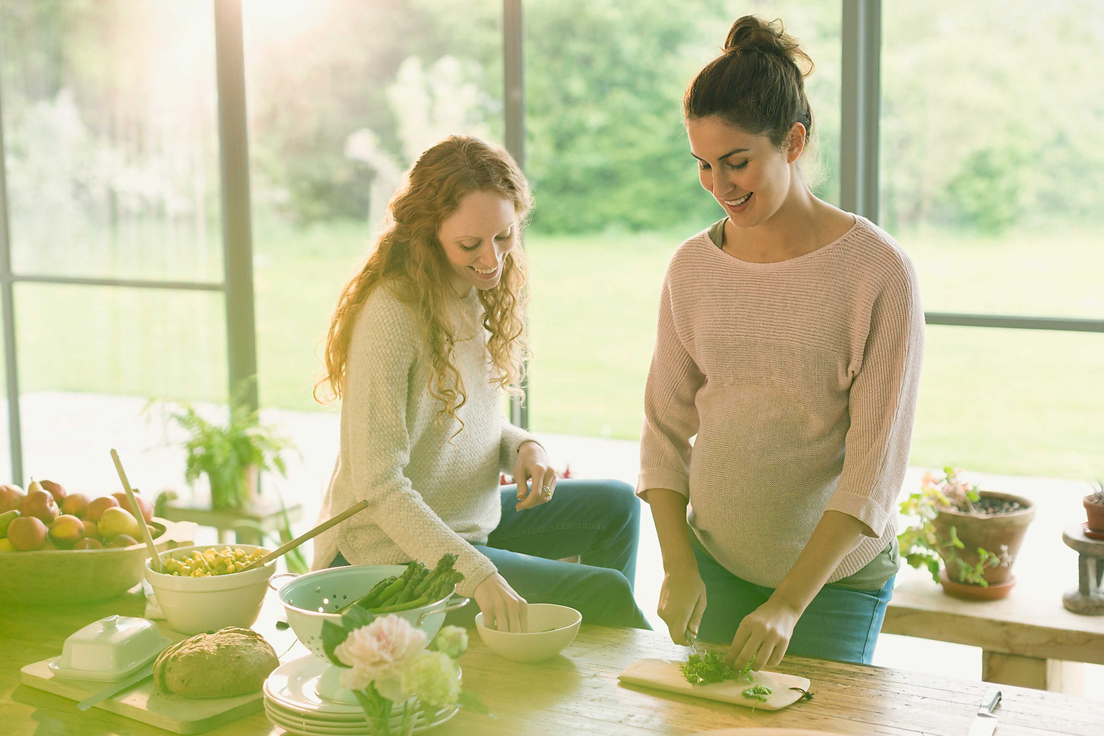 Schwangere Frau und Freundin bereiten in heller Küche frisches Gemüse zu und achten auf folatreiche Ernährung.
