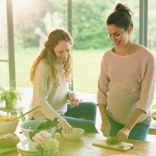Schwangere Frau und Freundin bereiten in heller Küche frisches Gemüse zu und achten auf folatreiche Ernährung.