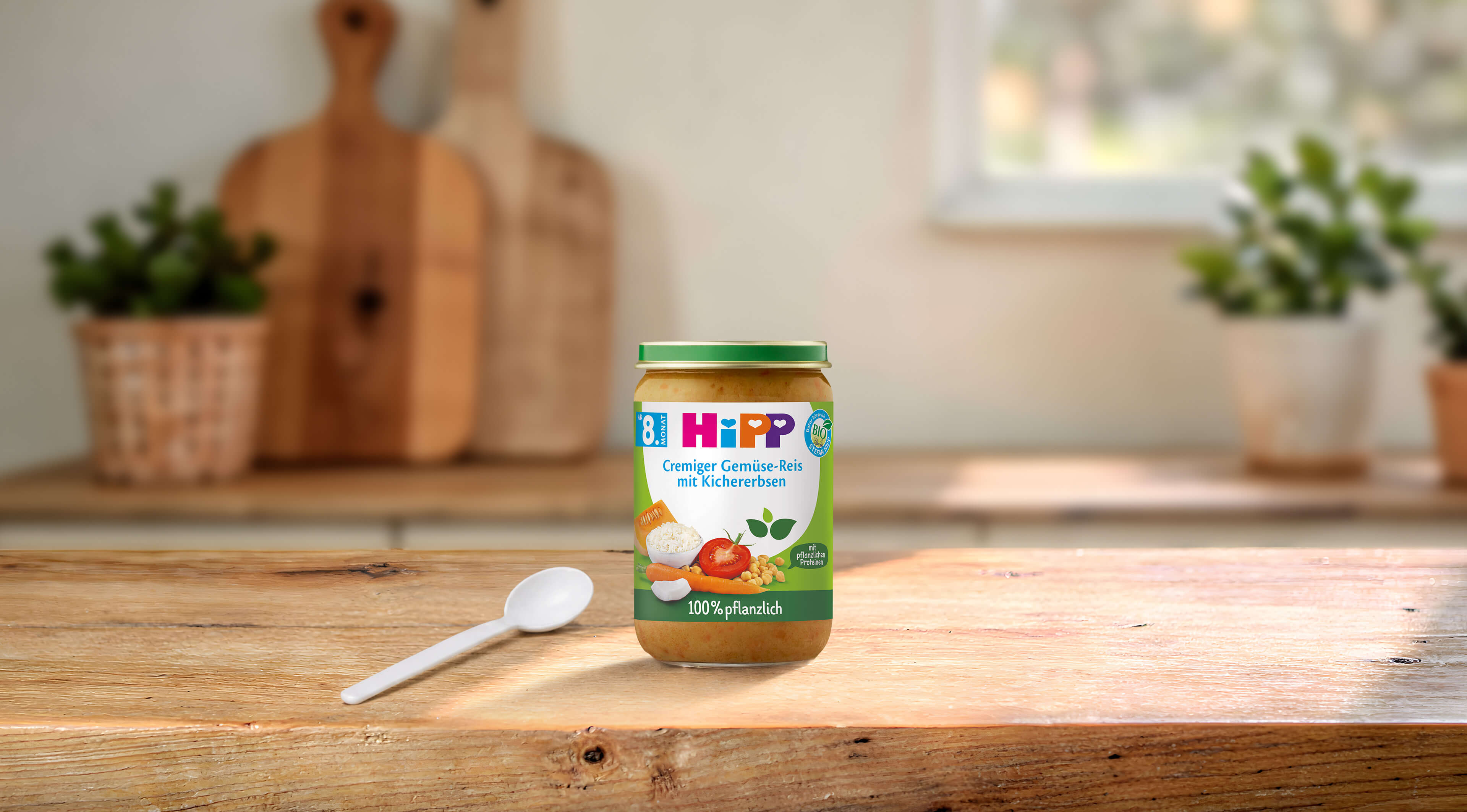 HiPP Bio-Gläschen „Cremiger Gemüse-Reis mit Kichererbsen“, 100 % pflanzlich, auf Holztisch mit Löffel in heller Küche