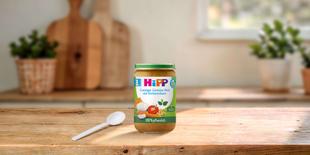 HiPP Bio-Gläschen „Cremiger Gemüse-Reis mit Kichererbsen“, 100 % pflanzlich, auf Holztisch mit Löffel in heller Küche