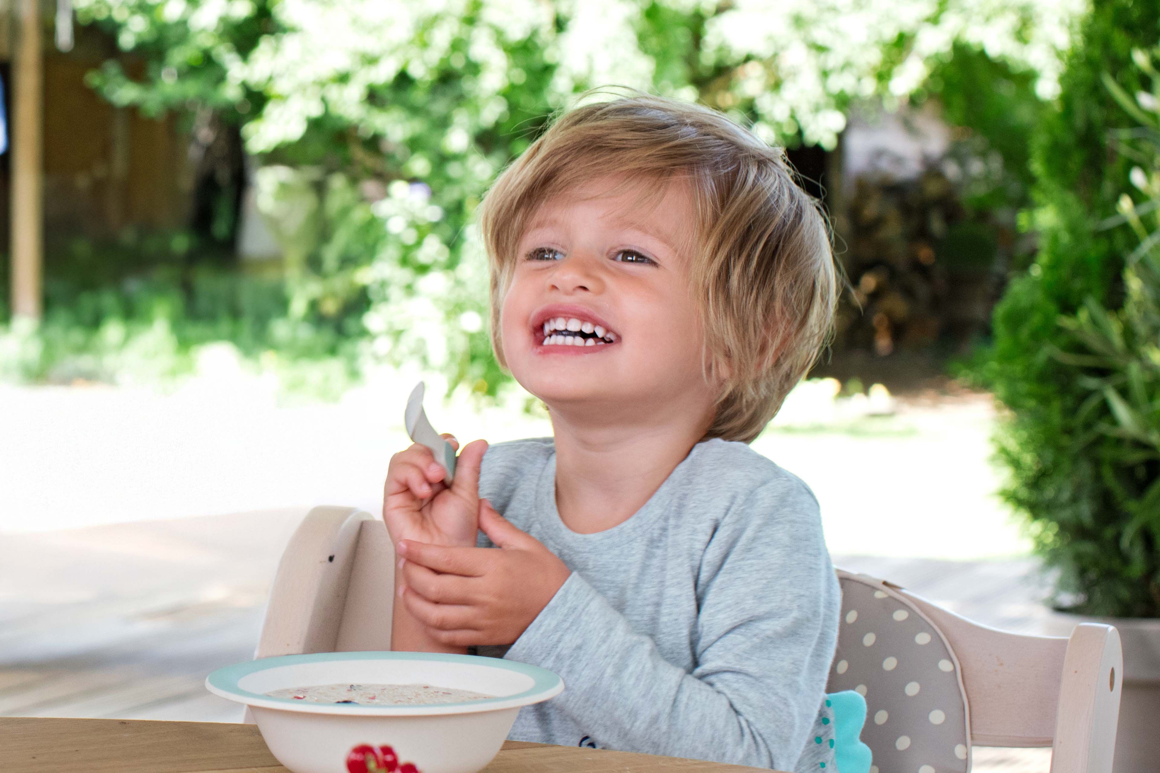 Lachendes Kleinkind sitzt draußen am Tisch und isst eine Schüssel Kindermüsli zum Frühstück.
