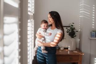 A mother wearing a white t-shirt and jeans holding her baby in a cosy grey knitted sleepsuit, looking out of a sunlit window.