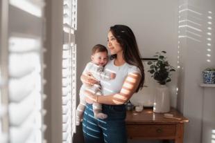 A mother wearing a white t-shirt and jeans holding her baby in a cosy grey knitted sleepsuit, looking out of a sunlit window.