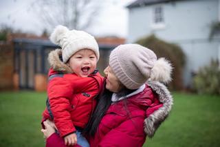 Woman holding a toddler in her arms outdoors both wearing a red coat and hats, the toddler is smiling and seems happy