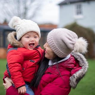 woman holding a toddler in her arms outdoors both wearing a red coat and hats, the toddler is smiling and seems happy.