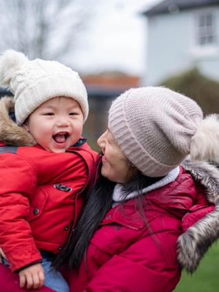 woman holding a toddler in her arms outdoors both wearing a red coat and hats, the toddler is smiling and seems happy.