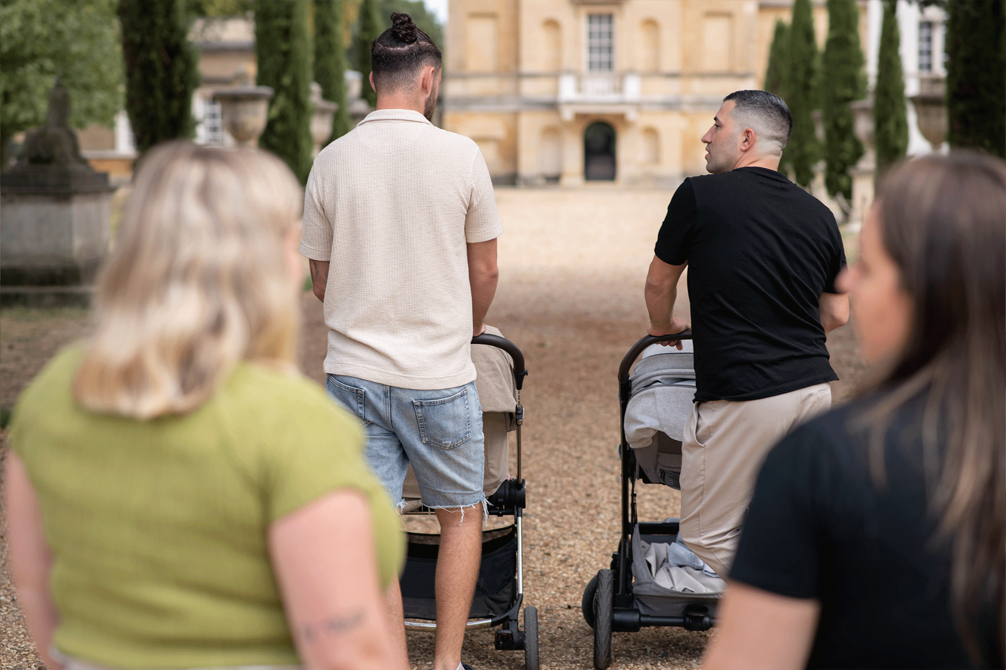 Four adults walk down a gravel path toward a large, historic stone building in the background. Two men lead the way, each pushing a stroller—one in a cream polo and denim shorts, and the other in a black t-shirt turned slightly to look back. In the foreground, two women walk alongside them, seen from behind.