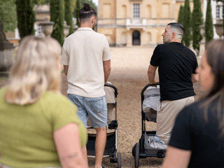 Four adults walk down a gravel path toward a large, historic stone building in the background. Two men lead the way, each pushing a stroller—one in a cream polo and denim shorts, and the other in a black t-shirt turned slightly to look back. In the foreground, two women walk alongside them, seen from behind.