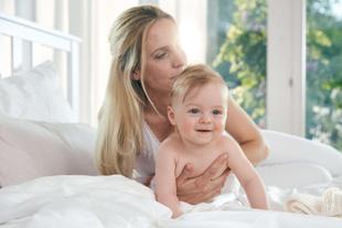 Mutter hält ihr nacktes Baby auf dem Bett, küsst es liebevoll im hellen Schlafzimmer mit Blick ins Grüne