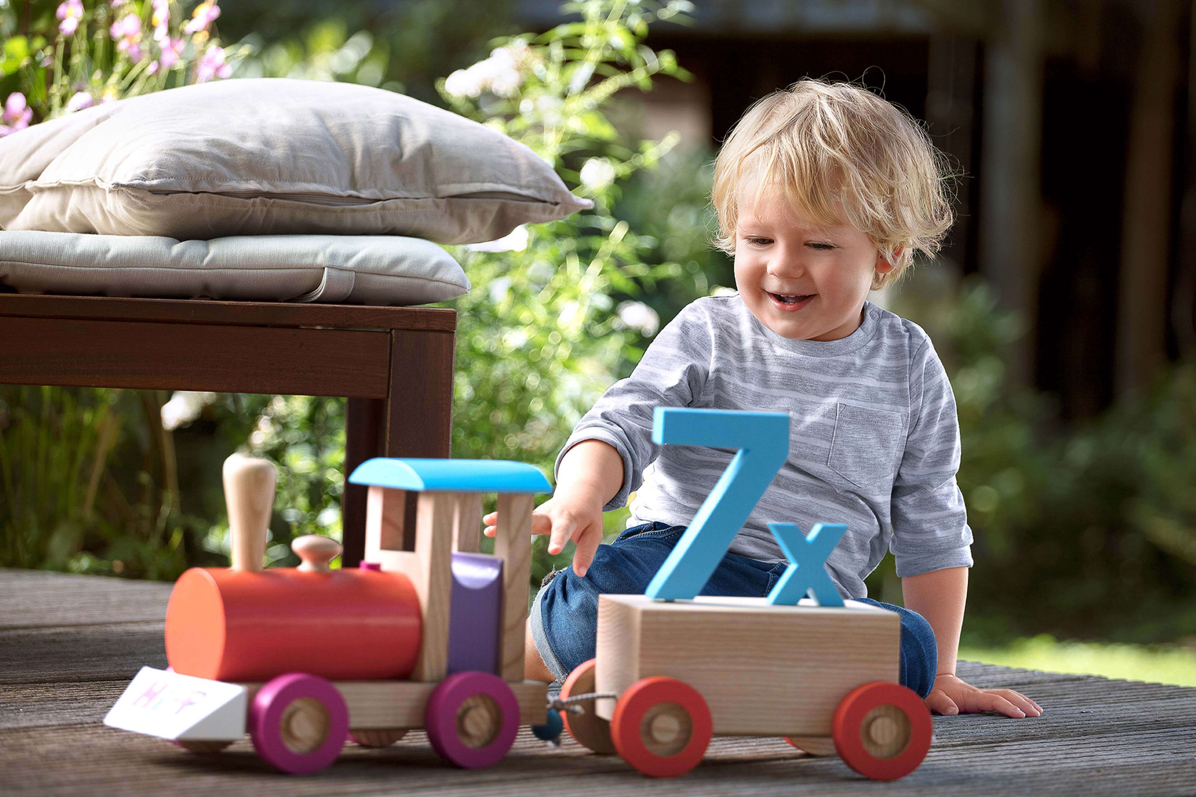 Lachendes Kleinkind spielt draußen mit einer Holzlok, auf deren Wagen eine große blaue „7x“ für Vitamin‑D‑Bedarf steht.