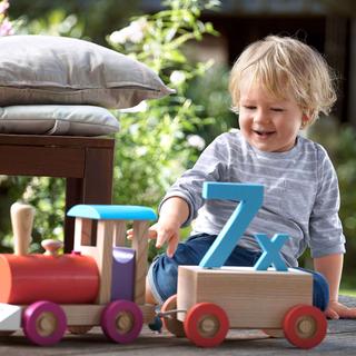 Lachendes Kleinkind spielt draußen mit einer Holzlok, auf deren Wagen eine große blaue „7x“ für Vitamin‑D‑Bedarf steht.