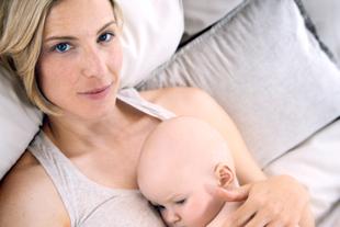Eine Frau mit blondem Haar und blauen Augen, die ein Baby an sich drückt, das auf einer weichen Unterlage liegt.