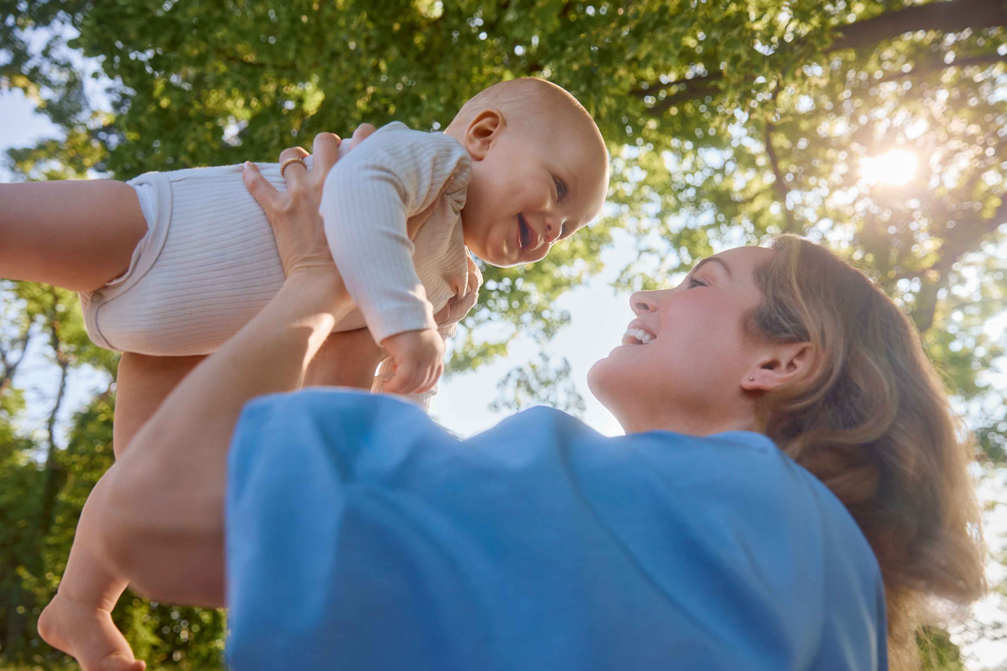Frau hebt lachendes Baby im Freien vor sonnigem Himmel