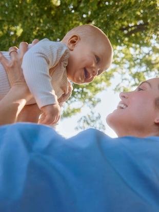 Frau hebt lachendes Baby im Freien vor sonnigem Himmel