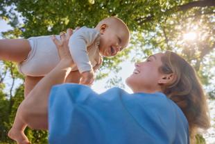 Lachende Mutter hebt ihr Baby im Park in die Luft, beide schauen sich liebevoll an, Sonnenlicht fällt durch die Bäume.