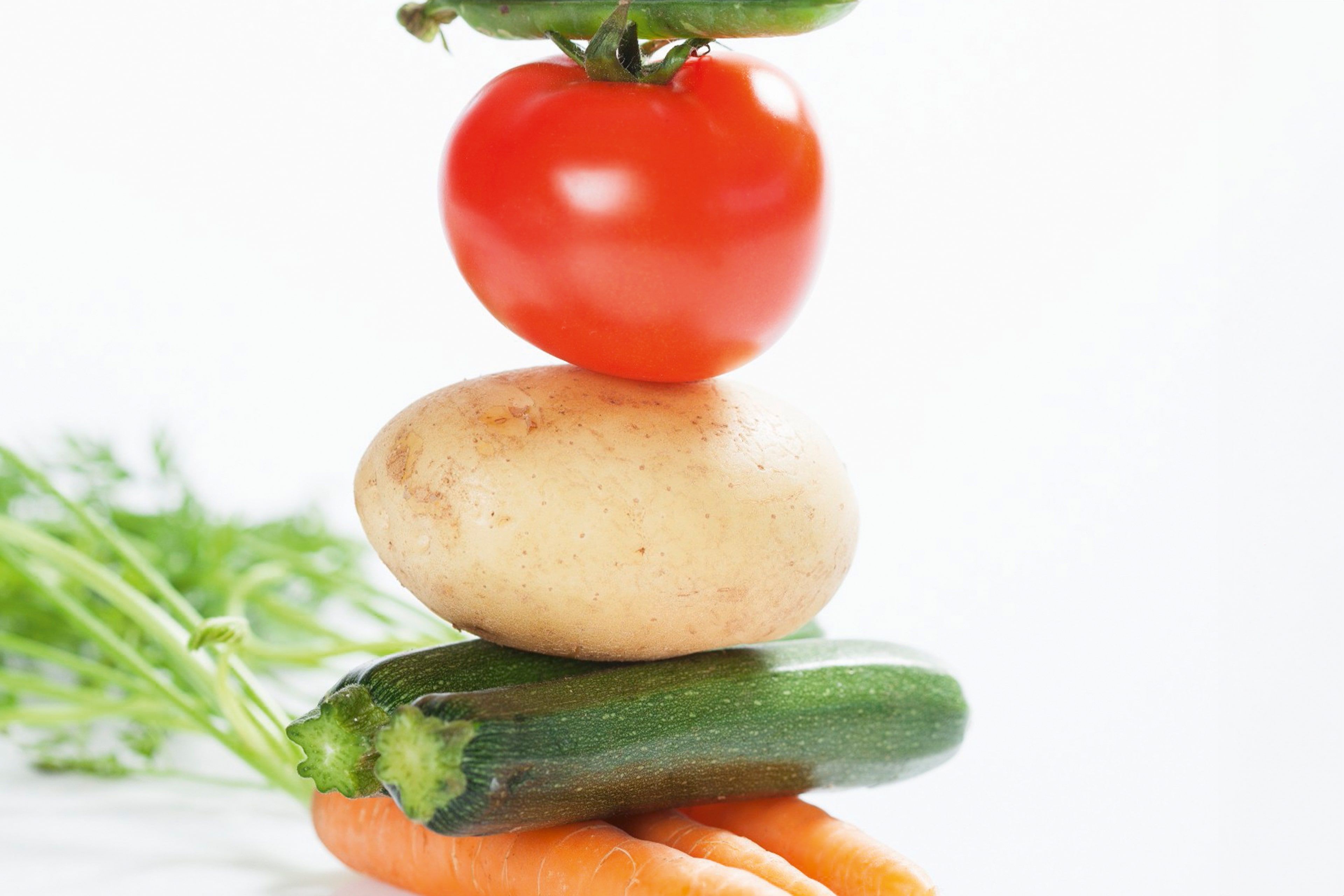 Ein Stapel frisches Gemüse, darunter eine Tomate, eine Kartoffel, eine Zucchini und eine Karotte, mit einem Hauch von grünen Kräutern im Hintergrund.