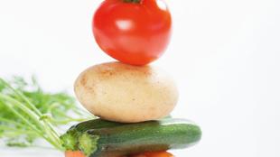 Ein Stapel frisches Gemüse, darunter eine Tomate, eine Kartoffel, eine Zucchini und eine Karotte, mit einem Hauch von grünen Kräutern im Hintergrund.
