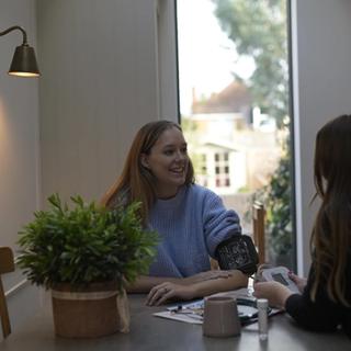 Pregnant woman smiling while having her blood pressure taken by a healthcare professional at a dining table in a bright, cozy home setting.