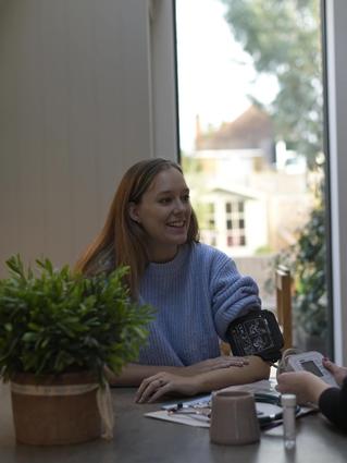 Pregnant woman smiling while having her blood pressure taken by a healthcare professional at a dining table in a bright, cozy home setting.