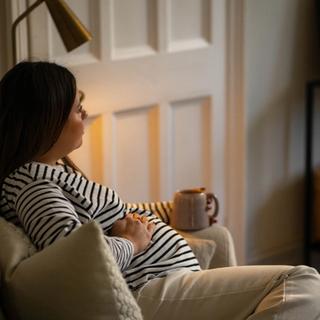 Pregnant woman relaxing on a couch with her hands on her belly, holding a mug, and gazing thoughtfully into the distance in a cozy living room.