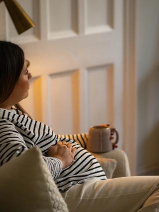 Pregnant woman relaxing on a couch with her hands on her belly, holding a mug, and gazing thoughtfully into the distance in a cozy living room.