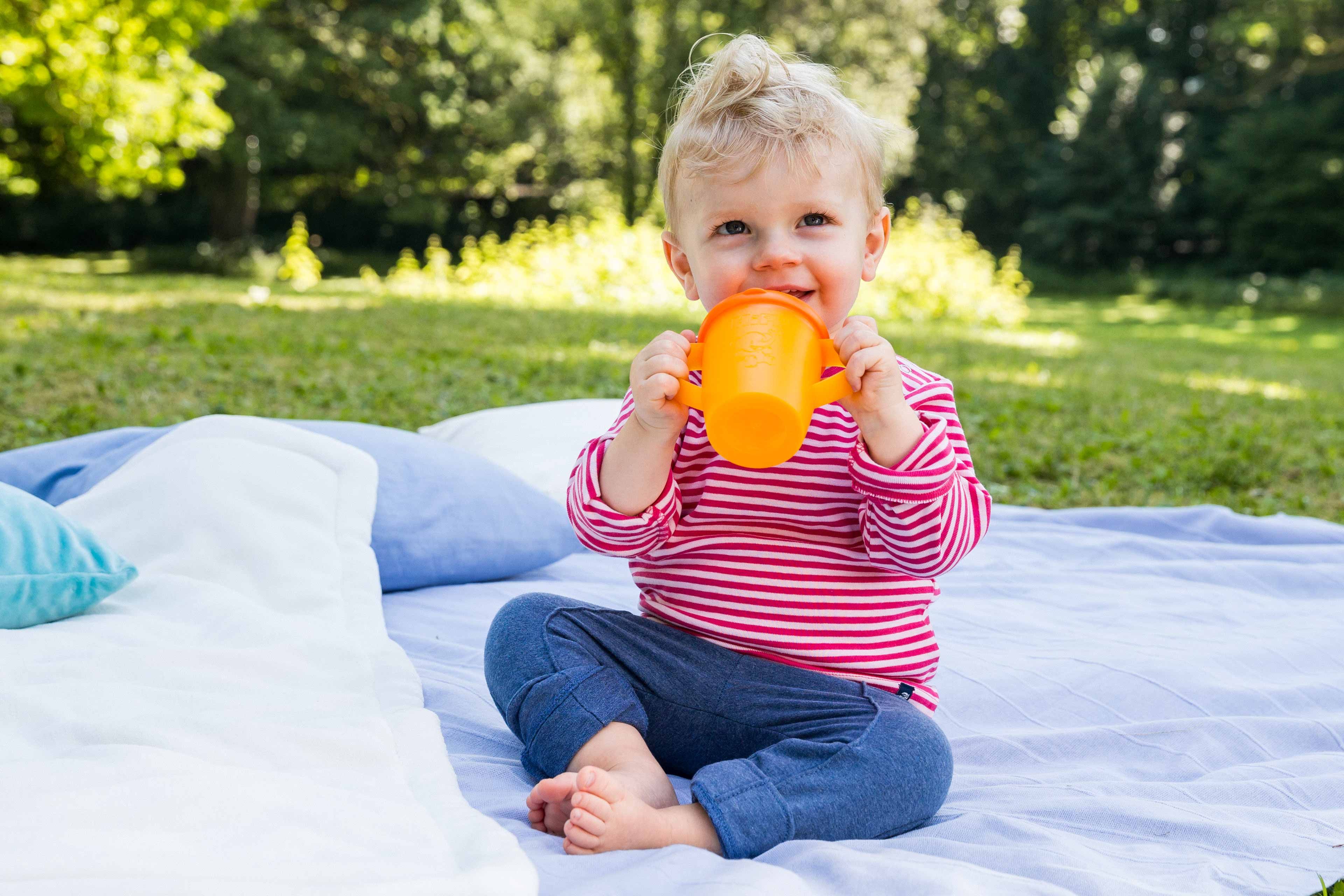 Ein kleines Kind, vermutlich ein Kleinkind, sitzt im Freien auf einer Decke und nippt aus einer orangefarbenen Schnabeltasse.