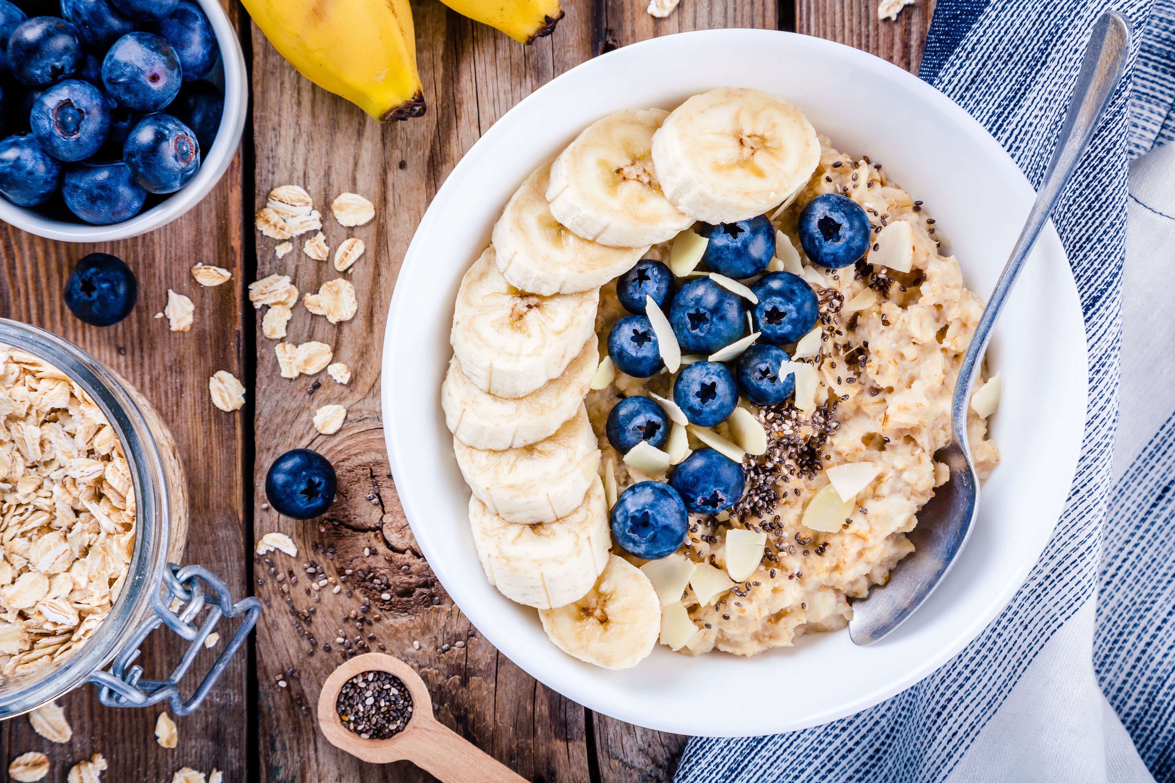 Eine Schüssel Haferflocken mit geschnittenen Bananen, Blaubeeren und Chiasamen, dazu ein Glas Haferflocken und eine Banane.
