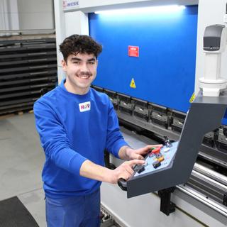 Lächelnder HiPP‑Auszubildender zum Industriemechaniker bedient eine große CNC‑Maschine in der Werkhalle.