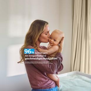 Frau hält lächelnd ein Baby im Arm und küsst es auf die Wange in einem hellen Raum mit Vorhängen